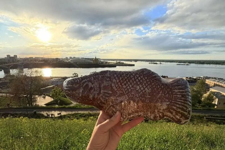 Большой пряник-щука в руке на фоне Волги и заречной части Нижнего Новгорода